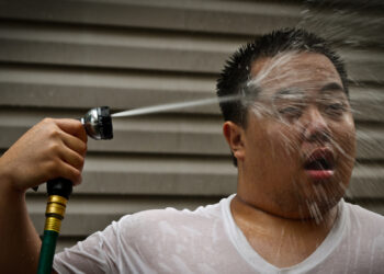 A man seeks relief during a heat wave with a garden hose shower. (Instant Vantage, CC BY-SA 2.0, via Wikimedia Commons)