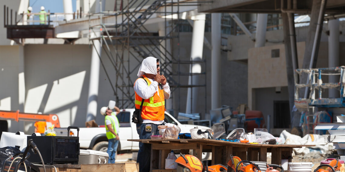 A construction worker ties a towel around his head while another worker can be seen drinking water from a gallon jug on a hot day. (iStock image)