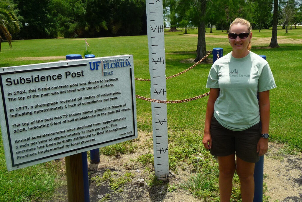 UCF Biology Associate Professor Lisa Chambers stands next to a subsidence post that marks soil subsidence in the Everglades Agricultural Area. Soil elevation has dropped as much as six feet over the past 100 years in some locations. (UCF photo)