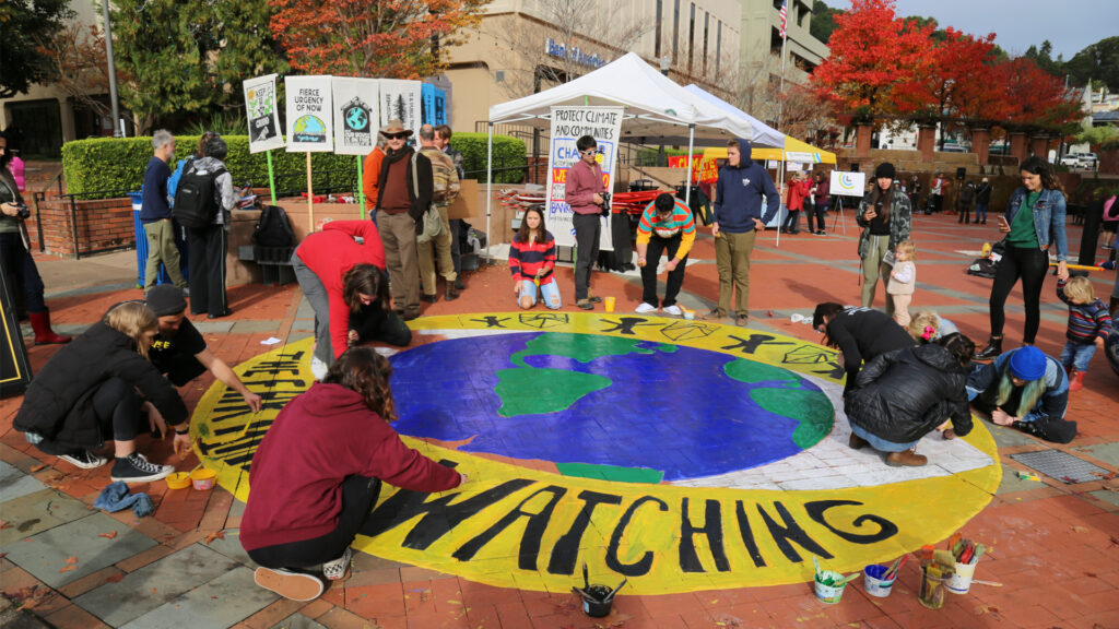 Members of the Sunrise Movement join in painting a mural reminding passers-by that “the earth is watching” before a climate protest in California. (Fabrice Florin, CC-BY-SA 3.0, via flickr)