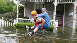 A National Guardsman giving a woman a piggyback in waist-deep floodwaters outside a house as part of rescue operations in flooded areas around Houston in 2017. (Zachary West/National Guard, CC BY 2.0, via Wikimedia Commons)