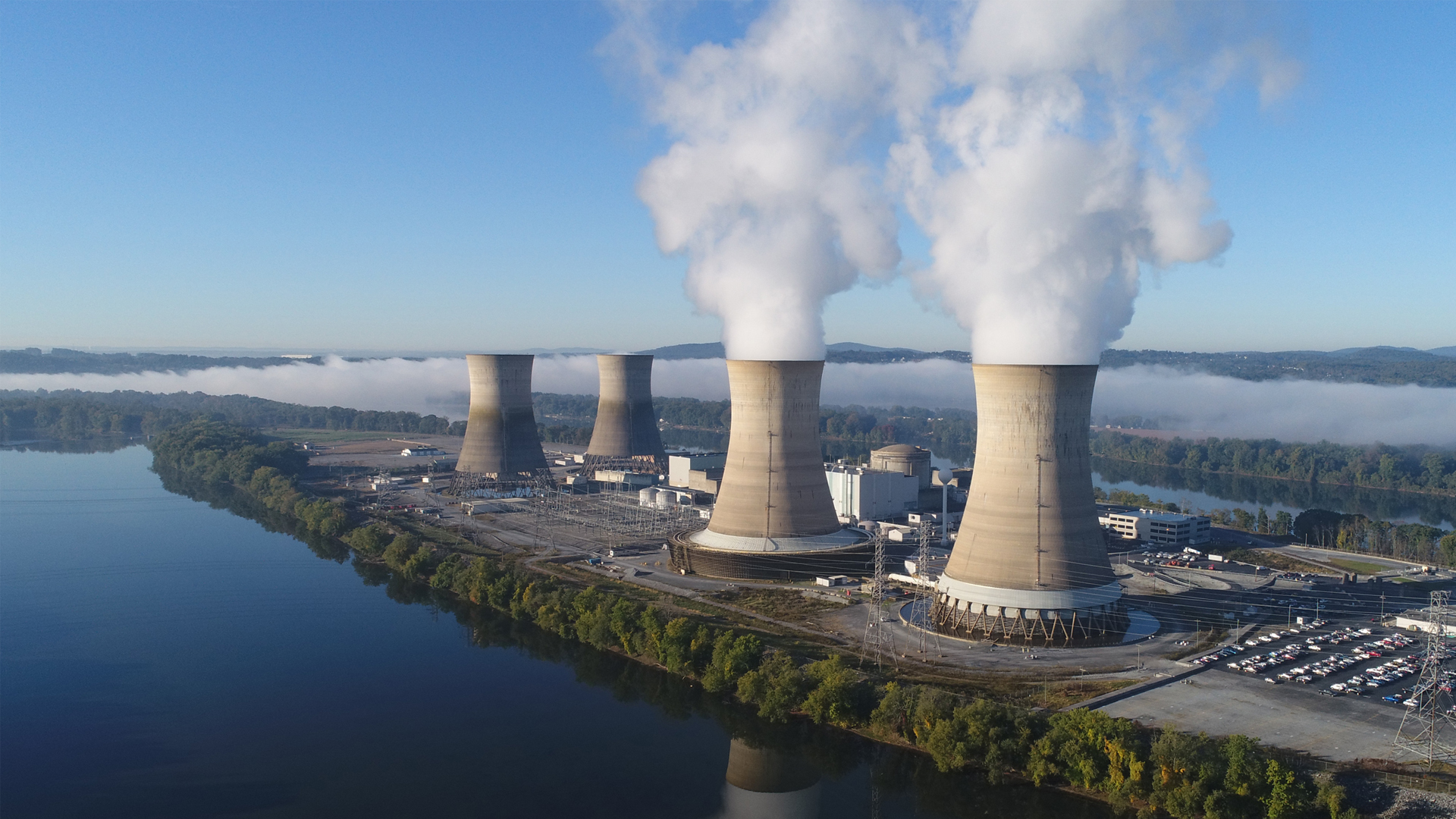 The Three Mile Island nuclear plant in 2019. The working cooling towers in the foreground are emitting water vapor. The dormant cooling towers are from Unit 2, which was permanently damaged in the 1979 accident. (Constellation Energy, CC BY-SA 4.0, via Wikimedia Commons)
