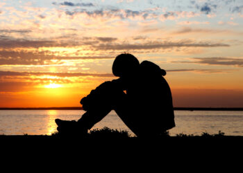 A young person sits by the water at sunset (iStock image)