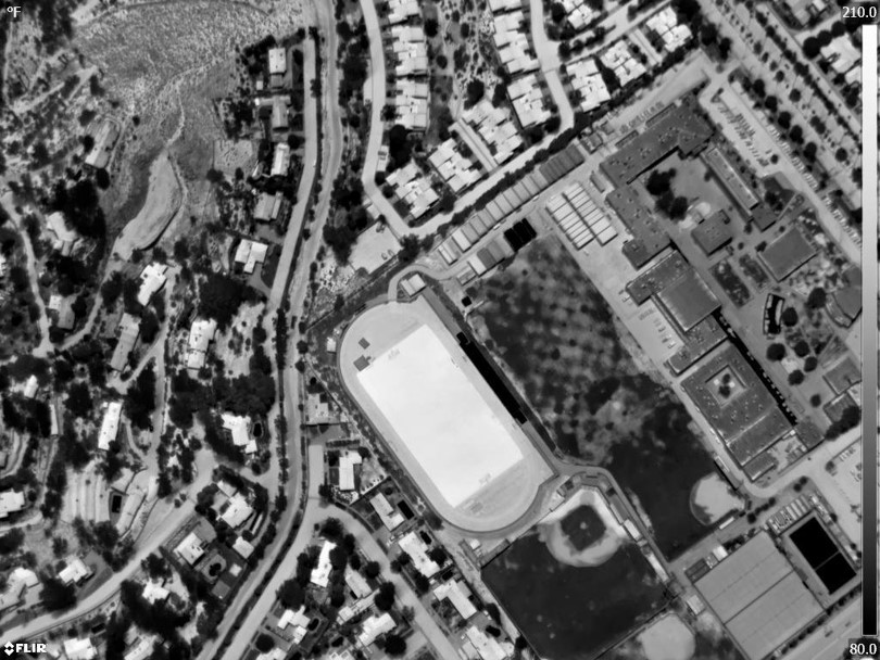 Aerial thermal image captured on August 8, 2019, shows a black and white view based on surface reflectance. At the center is a football stadium with artificial turf, adjacent to a baseball stadium with natural turf. The temperature scale, which appears along the right side of the photo, displays ground temperatures from 80°F to 210°F. The artificial turf in that image is 210°F. For a comparison, Schiavon states that is hotter than the parking lot shown in the same image. (Image courtesy of Marco Schiavon)
